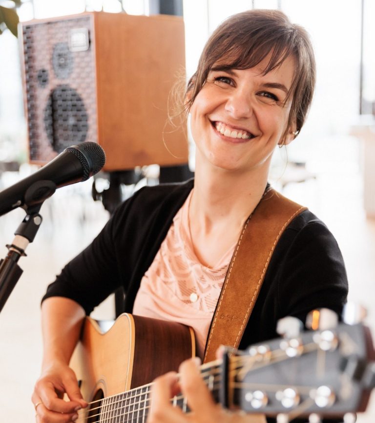 Bildnachweis: https://nd-lovestories.de/ Kate, spielbereit mit Mikrofon und im roten Blazer, fixiert ihren Capodaster an der Gitarre und blickt freundlich in die Kamera