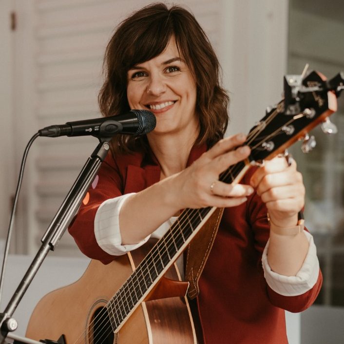 Kate, spielbereit mit Mikrofon und im roten Blazer, fixiert ihren Capodaster an der Gitarre und blickt freundlich in die Kamera